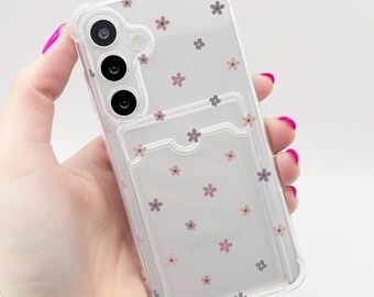 May include: Clear phone case with a floral pattern of small pink and gray flowers. The case has a cardholder on the back and is designed for a Samsung phone. The phone case is held in a hand with pink nail polish.