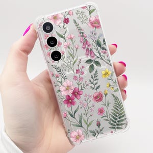 Puede incluir: Funda transparente para teléfono con un diseño floral de flores rosas, rojas y amarillas, hojas y tallos verdes. La funda tiene un borde transparente y tres recortes para la cámara.