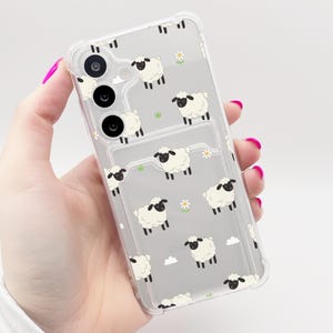 May include: A clear phone case featuring a repeating pattern of white sheep with black faces and legs, interspersed with small white daisies and green grass tufts. The case has reinforced corners and cutouts for the rear camera lenses.