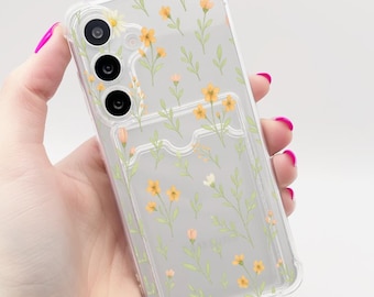 Puede incluir: Funda transparente para teléfono con un diseño floral de flores amarillas y blancas y hojas verdes. La funda tiene un tarjetero y se sostiene en una mano con esmalte de uñas rosa.