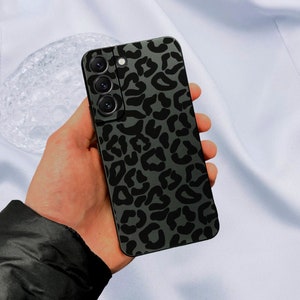 Puede incluir: Una funda de teléfono negra con un diseño de estampado de leopardo. La funda tiene una protuberancia de cámara elevada con tres lentes.
