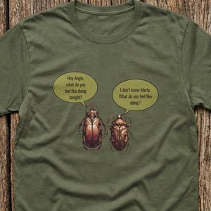 May include: Olive green t-shirt featuring a cartoon of two beetles with speech bubbles. The left beetle asks, "Hey Angie, what do you feel like doing tonight?" The right beetle replies, "I don't know Marty. What do you feel like doing?"