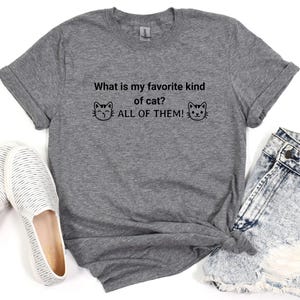 May include: Gray t-shirt with the text "What is my favorite kind of cat? ALL OF THEM!" and two cartoon cats on either side of the text.