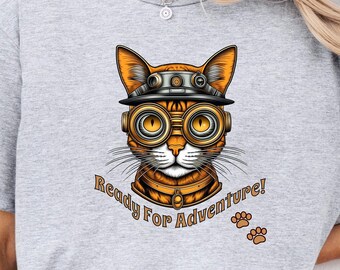 May include: A gray t-shirt with a cartoon illustration of an orange cat wearing goggles and a helmet. The text "Ready For Adventure!" is printed below the cat with two paw prints.