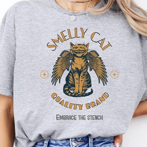 May include: Gray t-shirt with a graphic of a cat with wings. The text "Smelly Cat Quality Brand" is above the cat and "Embrace the Stench" is below the cat.