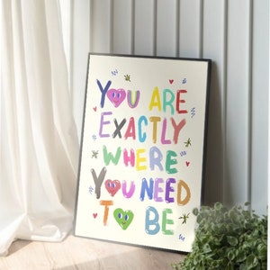 May include: A colorful framed print with the text "You are exactly where you need to be" in a playful, hand-drawn style. The print features hearts with smiley faces and stars.