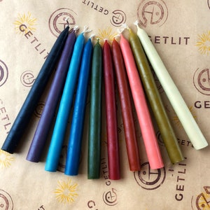 Puede incluir: Surtido de diez velas de tipo taper en varios colores, incluyendo negro, morado, azul, verde, rojo, marrón y crema. Las velas están dispuestas en forma de abanico sobre un fondo de papel marrón con el texto "GETLIT" impreso en el papel.