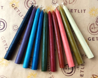 May include: Assortment of ten taper candles in various colours, including black, purple, blue, green, red, brown, and cream. The candles are arranged in a fan shape on a brown paper background with the text "GETLIT" printed on the paper.