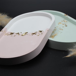 Könnte beinhalten: Zwei ovale Dekoschalen mit erhöhtem Rand. Die Schale links ist rosa mit einer weißen Oberseite und goldenen Flocken. Die Schale rechts ist hellgrün mit goldenen Flocken.