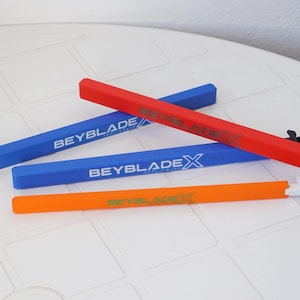 May include: Four colourful Beyblade X launchers in red, blue, and orange. The blue launchers have the text "BEYBLADE X" in white. The red launcher has black attachments on one end. The launchers are on a white surface.