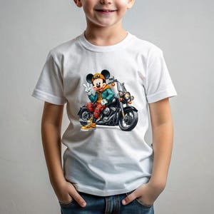 May include: White t-shirt with a graphic of Mickey Mouse riding a motorcycle. Mickey is wearing a blue jacket, red pants, and a yellow helmet. The motorcycle is black with chrome accents.