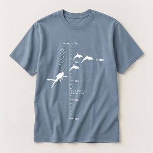 Könnte beinhalten: Ein staubblaues T-Shirt mit einer weißen Grafik eines Tauchers, Luftblasen und mehreren Orcas entlang einer vertikalen Tiefenskala von 0m bis 40m. Der Text "DIVE MORE WORRY LESS" ist in Weiß in der Nähe der 20m-Markierung aufgedruckt.