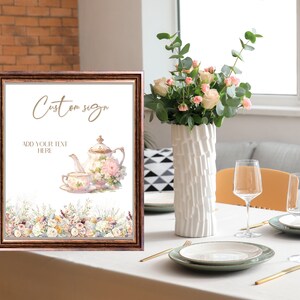 Op de afbeelding: Een printbaar bord met een bruin frame en een witte achtergrond. Het bord heeft een aquarel-illustratie van een theepot en een theekopje met bloemmotieven. De tekst "Custom sign" en "Add your text here" is in goud op het bord gedrukt.