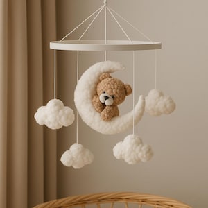 May include: A baby mobile featuring a brown teddy bear nestled in a white crescent moon, surrounded by fluffy white clouds. The mobile hangs from a white circular frame. A wicker bassinet is in the foreground.