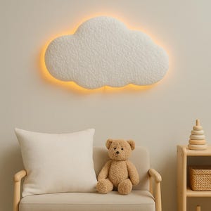 Könnte beinhalten: Ein wolkenförmiges Wandlicht mit warmem Schein erhellt ein neutrales Kinderzimmer. Ein beiger Teddybär sitzt auf einem kleinen Sofa mit einem cremefarbenen Kissen. Ein Holzregal hält einen Korb und Holzringe.