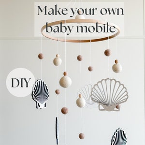 Peut inclure: Mobile bébé DIY avec un anneau en bois, des boules de feutre blanches et brunes et des coquillages en bois. Le texte "Make your own baby mobile" est en haut de l'image.