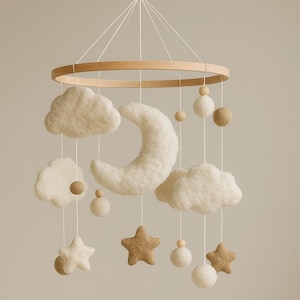 Könnte beinhalten: Baby-Mobile mit Holzring und Filz-Wolken, Sternen, Mond und Kugeln in Creme- und Beigetönen. Das Mobile hängt über einem weißen Babybett mit wolkenförmigen Kissen.
