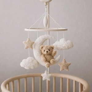 Può includere: Mobile per neonati beige con un orsacchiotto su una luna crescente, stelle e nuvole. Il mobile è appeso sopra una culla in legno. Design morbido e neutro, adatto a una cameretta.