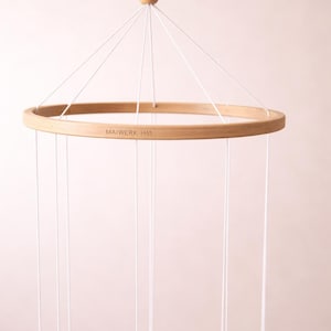 Può includere: Un mobile per bambini in legno con una cornice circolare e fili sospesi. La cornice è in legno chiaro e i fili sono bianchi. Il mobile è sospeso da una singola perla e corda di legno. Il testo "MAIWERK H&S" è stampato sulla cornice.