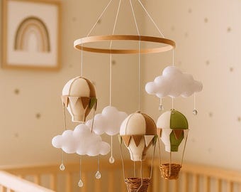 May include: A baby mobile featuring three hot air balloons in cream, brown, and green, suspended from a wooden ring. White felt clouds and dangling crystal beads add to the nursery decor. The mobile hangs above a wooden crib.