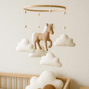 May include: A baby mobile featuring a tan horse with a white mane and tail, suspended below a wooden ring. White felt clouds are positioned around the horse. The mobile hangs above a wooden crib. Additional cloud-shaped pillows are in the crib.