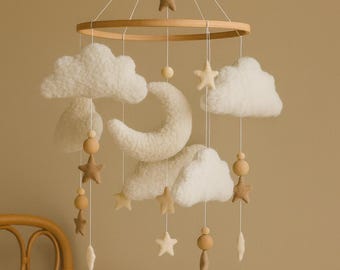 Op de afbeelding: Een baby mobile met een houten ring met hangende vilten wolken, sterren en een halve maan. De mobile is opgehangen aan witte touwtjes en bevat houten kralen. Het kleurenpalet is neutraal, met crème, beige en bruine tinten.