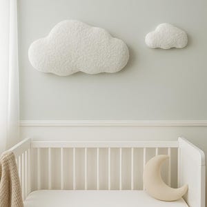 May include: A nursery scene featuring a white crib with a fitted sheet and a crescent moon pillow. Two fluffy, white cloud-shaped wall decorations are mounted on the light gray wall. A tan blanket hangs over the side of the crib.