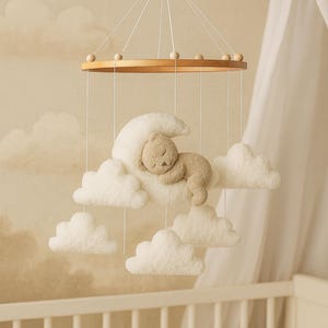 Puede incluir: Móvil para bebé con un anillo de madera, cuerdas, una luna de peluche, nubes y un osito de peluche durmiendo. El móvil está suspendido sobre una cuna blanca, creando una decoración suave y soñadora para la guardería.