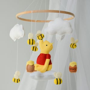 May include: A Winnie the Pooh themed baby mobile with a yellow felt Winnie the Pooh sitting on a white cloud, surrounded by yellow and black felt bees and white clouds. The mobile is hanging from a wooden ring.