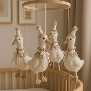Pode incluir: Um móbile para bebé com quatro gansos de pelúcia. Cada ganso é creme com bico e patas bege, usando um chapéu branco com pompom e uma gola bege com uma fita branca. O móbile está pendurado sobre um berço de madeira.