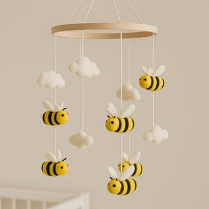 Może przedstawiać: Karuzela dla dziecka z filcowymi pszczołami i chmurkami. Pszczoły są żółte z czarnymi paskami i białymi skrzydłami. Chmury są białe. Karuzela zawieszona jest na drewnianym kole za pomocą białych sznurków.