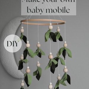 May include: DIY baby mobile with a wooden ring and green felt leaves hanging from white strings with wooden beads. The mobile is hanging from the ceiling.
