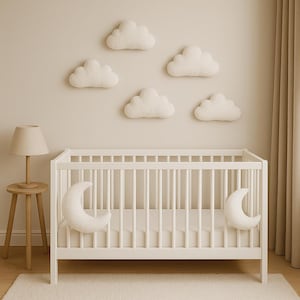 May include: A white cot with a mattress, and two crescent moon pillows. Five fluffy cloud-shaped wall decorations are above the cot. A small wooden side table with a lamp is to the left. The room has a neutral colour palette.