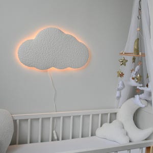 Könnte beinhalten: Ein weißes, wolkenförmiges Nachtlicht mit warmem Schein beleuchtet ein weißes Babybett mit einem weißen und goldenen Mobile, das darüber hängt.