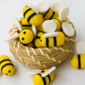 Può includere: Un gruppo di api in feltro fatte a mano in un cestino intrecciato. Le api sono gialle con strisce nere e ali bianche. Alcune api sono sparse intorno al cestino. Il cestino è fatto di materiale intrecciato marrone chiaro.