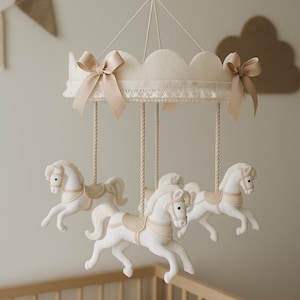 Può includere: Mobile a carosello color crema con tre cavalli bianchi, ciascuno con una sella beige. Il mobile ha una parte superiore smerlata con fiocchi beige ed è sospeso da una corda. Sotto è visibile una culla di legno.
