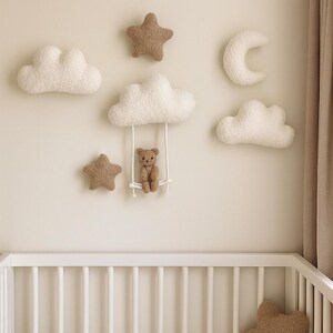 May include: A nursery wall decoration set with fluffy white clouds, brown stars, and a crescent moon. A small teddy bear sits on a swing hanging from a cloud. A teddy bear-shaped pillow rests in a white cot.