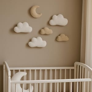 Op de afbeelding: Een kinderkamerscène met een witte wieg en wanddecoraties. De muur is voorzien van pluche wolken en maanvormen in wit en beige. De wieg heeft een wit frame en een witte matras, wat een zachte, dromerige esthetiek creëert.