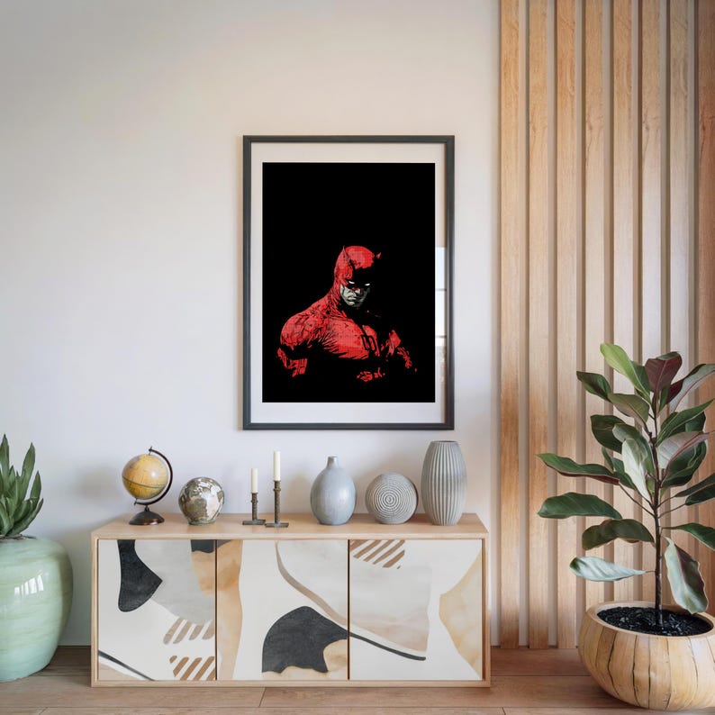 Pu&ograve; includere: Stampa artistica incorniciata con un'illustrazione rossa e nera di un supereroe. L'opera d'arte &egrave; appesa a una parete bianca sopra un mobile in legno con oggetti decorativi. Sono visibili anche una pianta in vaso e un mappamondo.