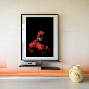 Pu&ograve; includere: Una stampa artistica incorniciata con un'illustrazione rossa e nera di un supereroe. L'opera raffigura una figura in costume rosso su sfondo nero. La stampa &egrave; esposta su una parete sopra una mensola con un piccolo componente audio.