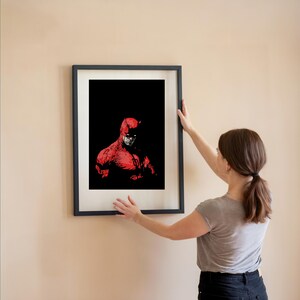 Pu&ograve; includere: Una stampa artistica incorniciata con un'illustrazione rossa e nera di un supereroe. L'opera raffigura un personaggio con un costume rosso e corna su sfondo nero. La stampa &egrave; in una cornice nera.