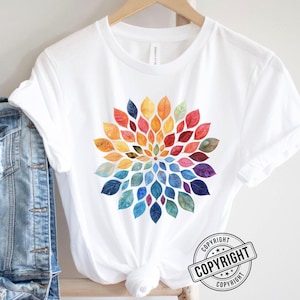 K&ouml;nnte beinhalten: Ein wei&szlig;es kurz&auml;rmeliges T-Shirt mit einer kreisf&ouml;rmigen Mandala-Grafik, die aus bunten Blattformen in Rot-, Orange-, Gelb-, Gr&uuml;n-, Blau- und Violettt&ouml;nen besteht. Das T-Shirt h&auml;ngt auf einem Holzkleiderb&uuml;gel neben einem blauen Jeansstoff.