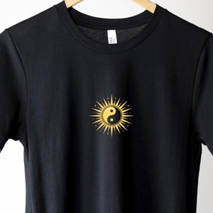 Könnte beinhalten: Schwarzes T-Shirt mit einem goldenen Sonnendesign und einem Yin-und-Yang-Symbol in der Mitte. Die Sonne hat spitze Strahlen. Das T-Shirt hängt an einem Holzbügel.