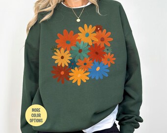 May include: A dark green sweatshirt featuring a floral design with orange, yellow, blue, and teal flowers. The sweatshirt has a crew neck and long sleeves. A yellow circle with the text "MORE COLOR OPTIONS" is in the lower left corner.