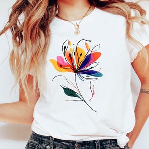 May include: White t-shirt featuring a vibrant, abstract floral design. The artwork showcases a colorful array of pink, yellow, orange, and blue hues, with black line art detailing the petals and stem. The shirt is a casual, everyday wear item.