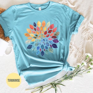 K&ouml;nnte beinhalten: Ein t&uuml;rkisfarbenes T-Shirt mit kurzen &Auml;rmeln und einer kreisf&ouml;rmigen Mandala-Grafik, die aus zahlreichen kleinen Bl&auml;ttern in einem Farbverlauf aus Rot, Orange, Gelb, Gr&uuml;n, Blau und Violett besteht. Das Shirt hat einen klassischen Rundhalsausschnitt.