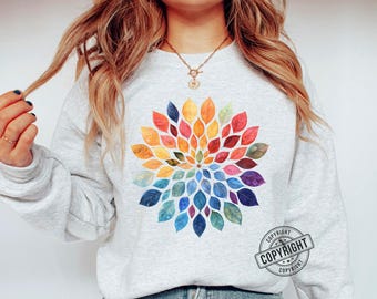 May include: A light gray sweatshirt featuring a colorful floral design. The design is composed of watercolor-style leaves in shades of red, orange, yellow, green, blue, and purple, arranged in a circular pattern. A copyright stamp is visible.