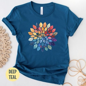 K&ouml;nnte beinhalten: Ein tiefblaugr&uuml;nes Kurzarm-T-Shirt mit einer zentralen kreisf&ouml;rmigen Grafik aus aquarellartigen Bl&auml;ttern in einem Farbspektrum aus Gelb, Orange, Rot, Violett und Blau. Das T-Shirt hat einen Rundhalsausschnitt und eine normale Passform.