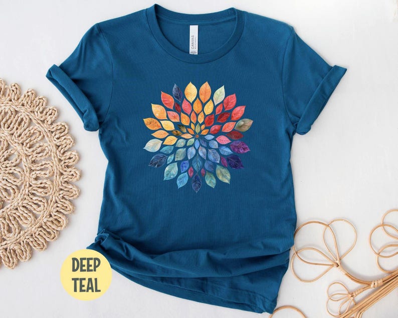 K&ouml;nnte beinhalten: Ein tiefblaugr&uuml;nes Kurzarm-T-Shirt mit einer zentralen kreisf&ouml;rmigen Grafik aus aquarellartigen Bl&auml;ttern in einem Farbspektrum aus Gelb, Orange, Rot, Violett und Blau. Das T-Shirt hat einen Rundhalsausschnitt und eine normale Passform.