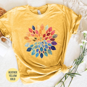 K&ouml;nnte beinhalten: Ein heather-gelbgoldenes T-Shirt mit einem kreisf&ouml;rmigen Grafikdesign, das aus aquarellartigen Bl&auml;ttern in einem Farbspektrum aus Rot, Orange, Gelb, Gr&uuml;n, Blau und Violett besteht, die in einem radialen Muster auf der Brust angeordnet sind.